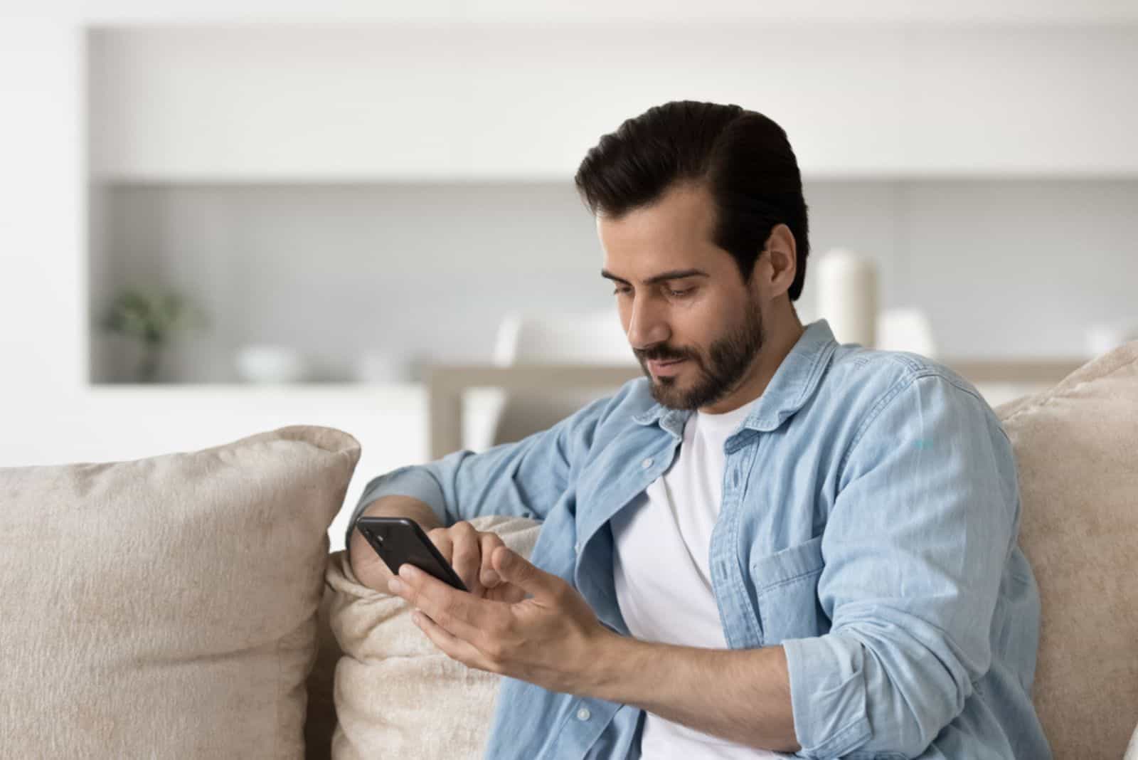 Mietteliäs vakava nuori mies istuu sohvalla kotona tekstiviestinä