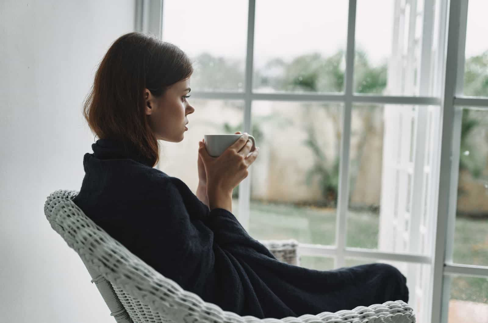 nainen istuu ikkunan vieressä juomassa kahvia